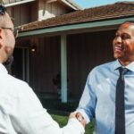Close-up of a professional handshake between a real estate agent and a client over a modern desk with a set of house keys,...