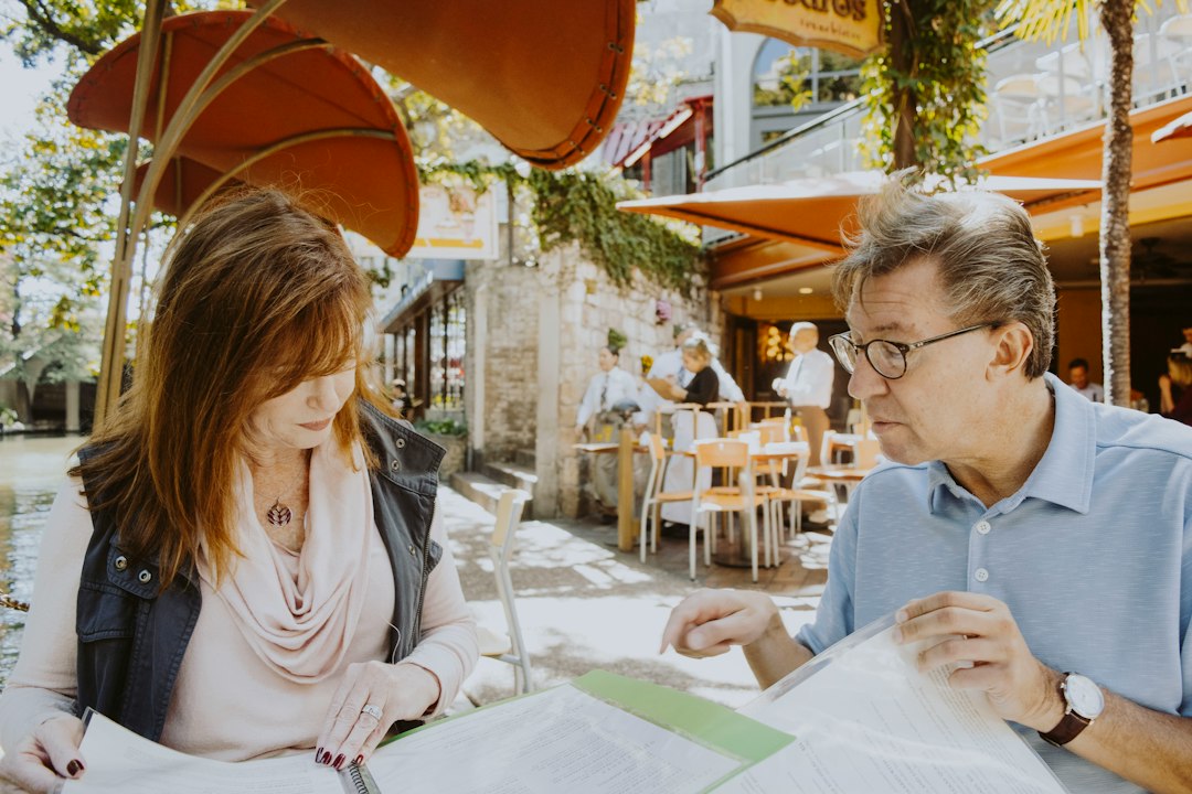 A senior couple sits at a sunlit kitchen table, looking thoughtfully at papers and a laptop, collaboratively planning their finances.
